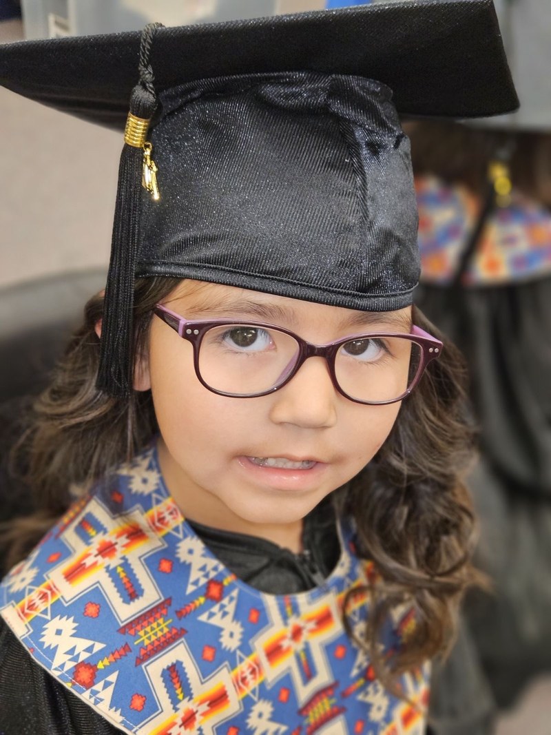 A young graduate in a black cap and gown with a brightly patterned stole, smiling at the camera. Another child in a cap stands behind.