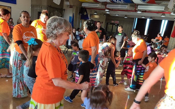 Staff and children holding hands in a round dance at an Orange Shirt Day gathering.