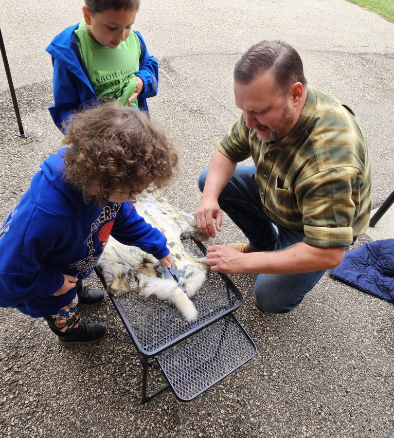 A child scrapes a stretched hide with a bone tool while a mentor steadies it.