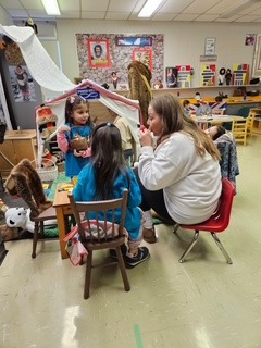 Children and a staff member in the classroom building a small shelter from a sheet and poles, with a hide and craft materials beside them.