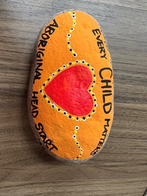 A smooth stone painted orange with a red heart at its centre, outlined in black, white and yellow dots. &ldquo;Every child matters&rdquo; is lettered around one side and &ldquo;Aboriginal Head Start&rdquo; up the other.