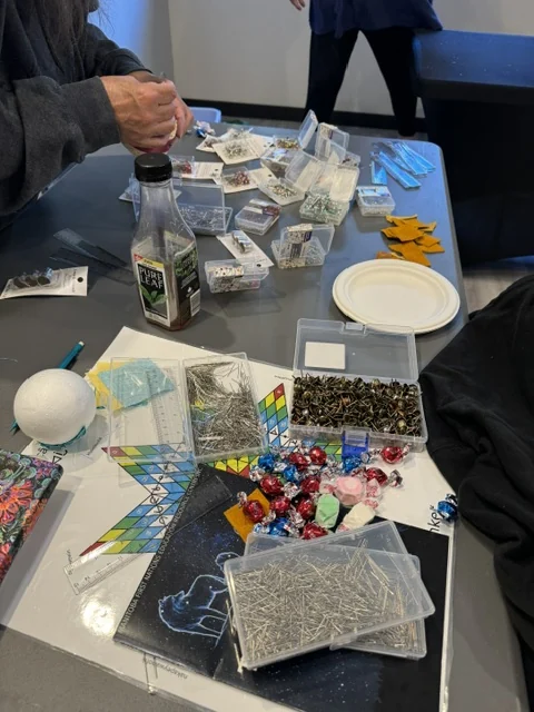 A work table laid out with supplies — boxes of pins, sequins and beads, a foam ball, a ruler and printed pattern cards — with a participant reaching across.