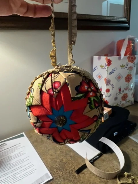 A red and blue ornament held up by its gold trim, with the work tables and supplies behind it.