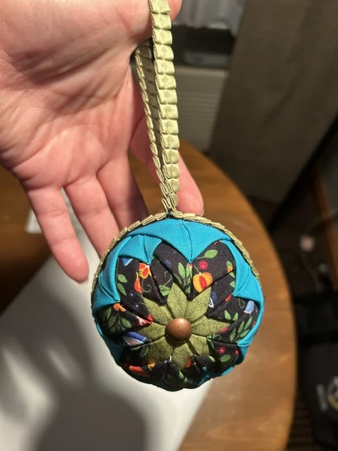 An ornament in olive green and patterned fabric resting on an open hand, its gold ribbon hanger trailing across the palm.