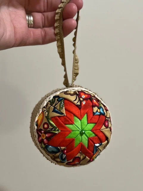 A finished red and green ornament hanging from a gold ribbon held between finger and thumb.