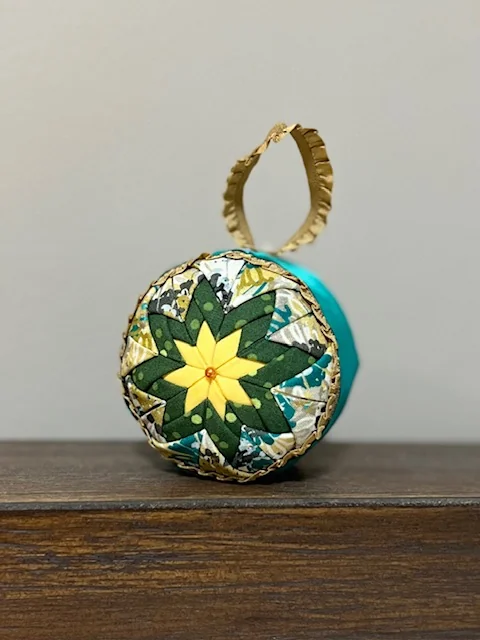 A teal, green and yellow ornament standing upright on a wooden table, its ribbon hanger looped above it.