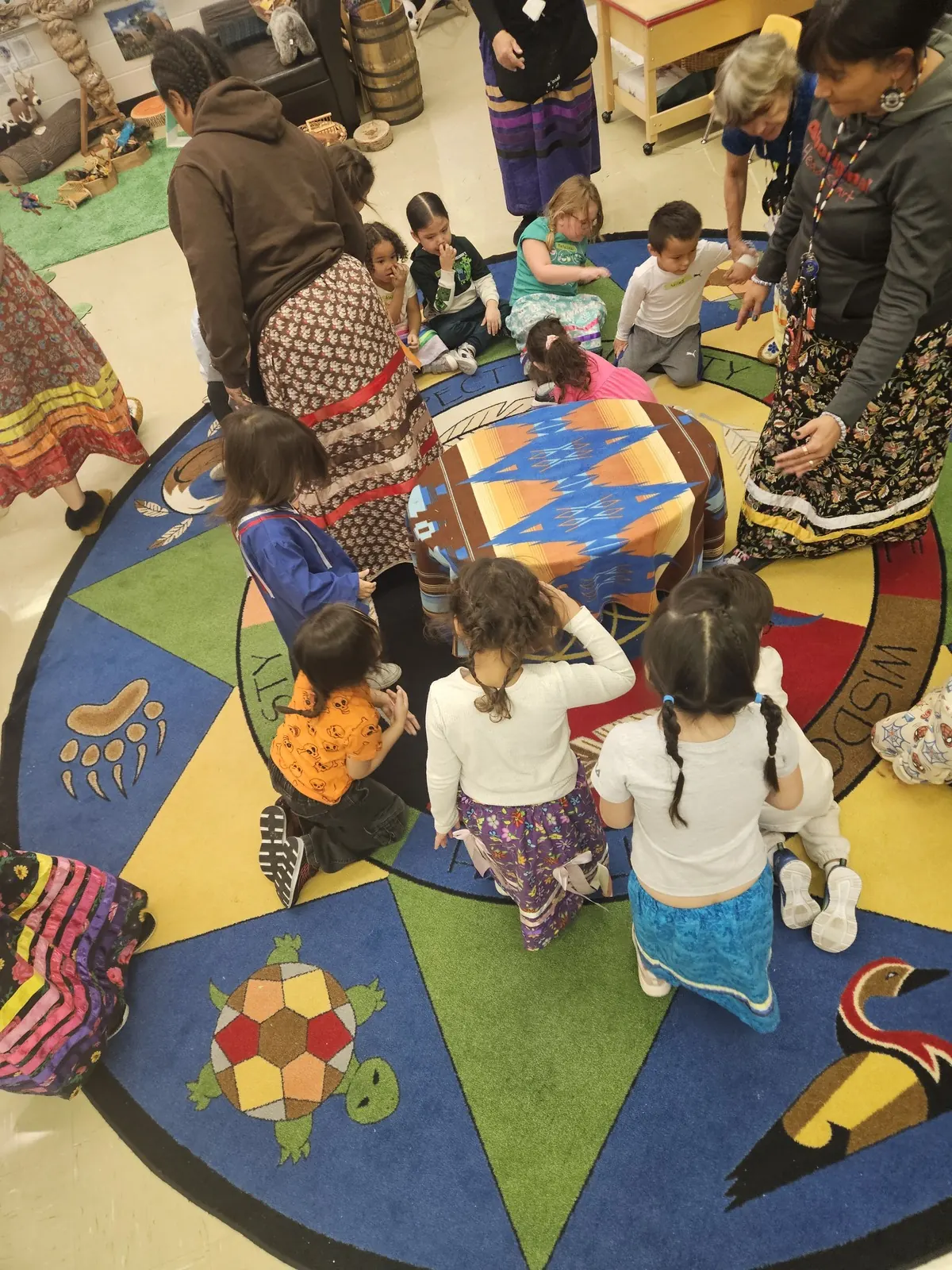 Children kneeling in a circle on the medicine wheel rug, seen from above.