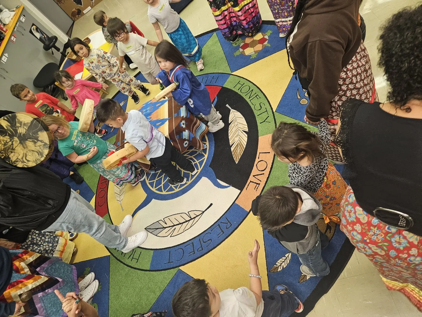 A large circle of children and adults gathered on the rug with drums in the middle.