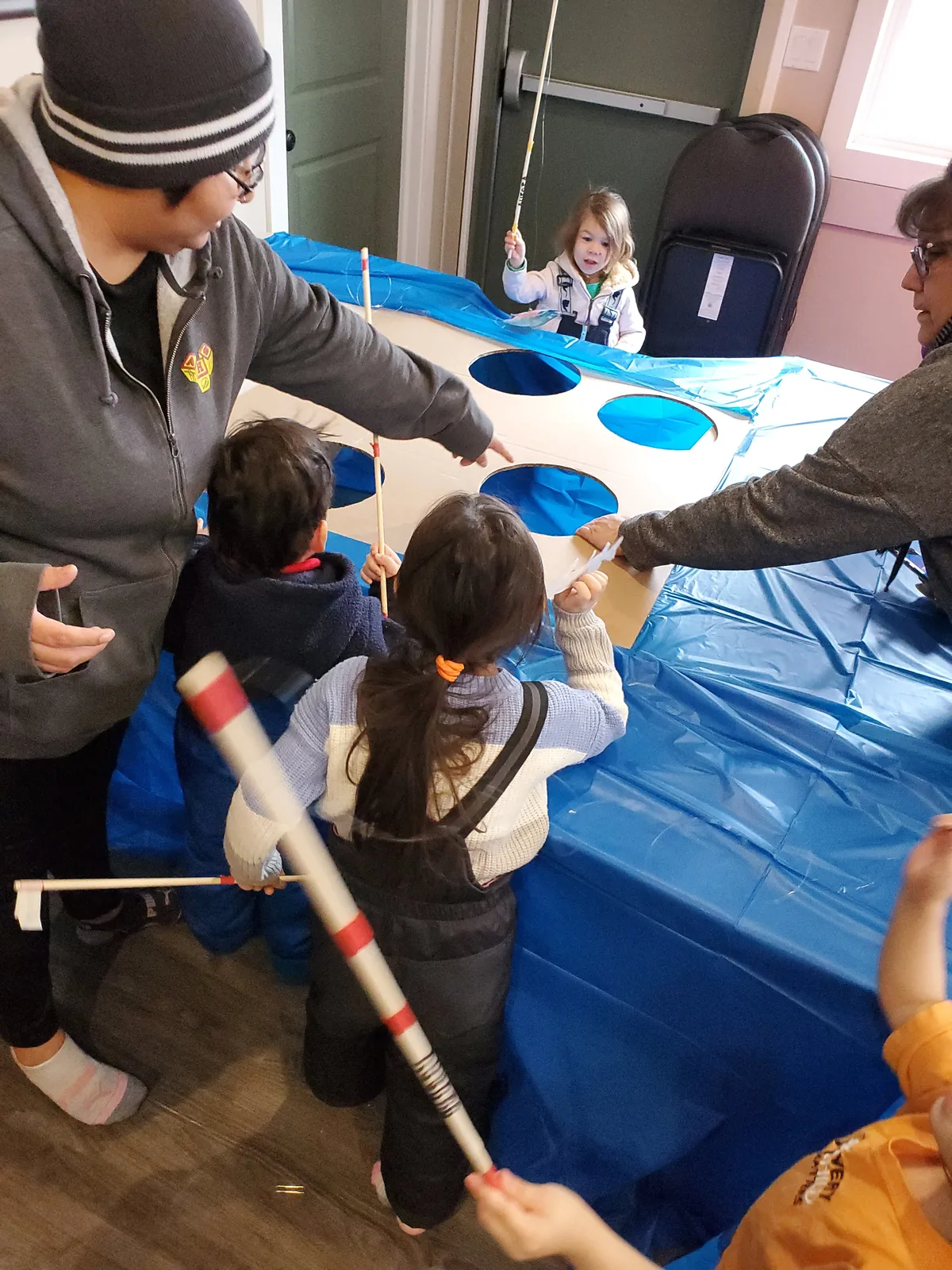Children practising with wooden rods through holes cut in a board laid over a blue tarp, standing in for the ice.