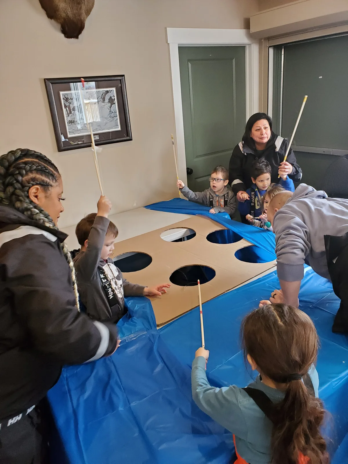 More children at the practice board, rods raised, an adult helping from the far side.