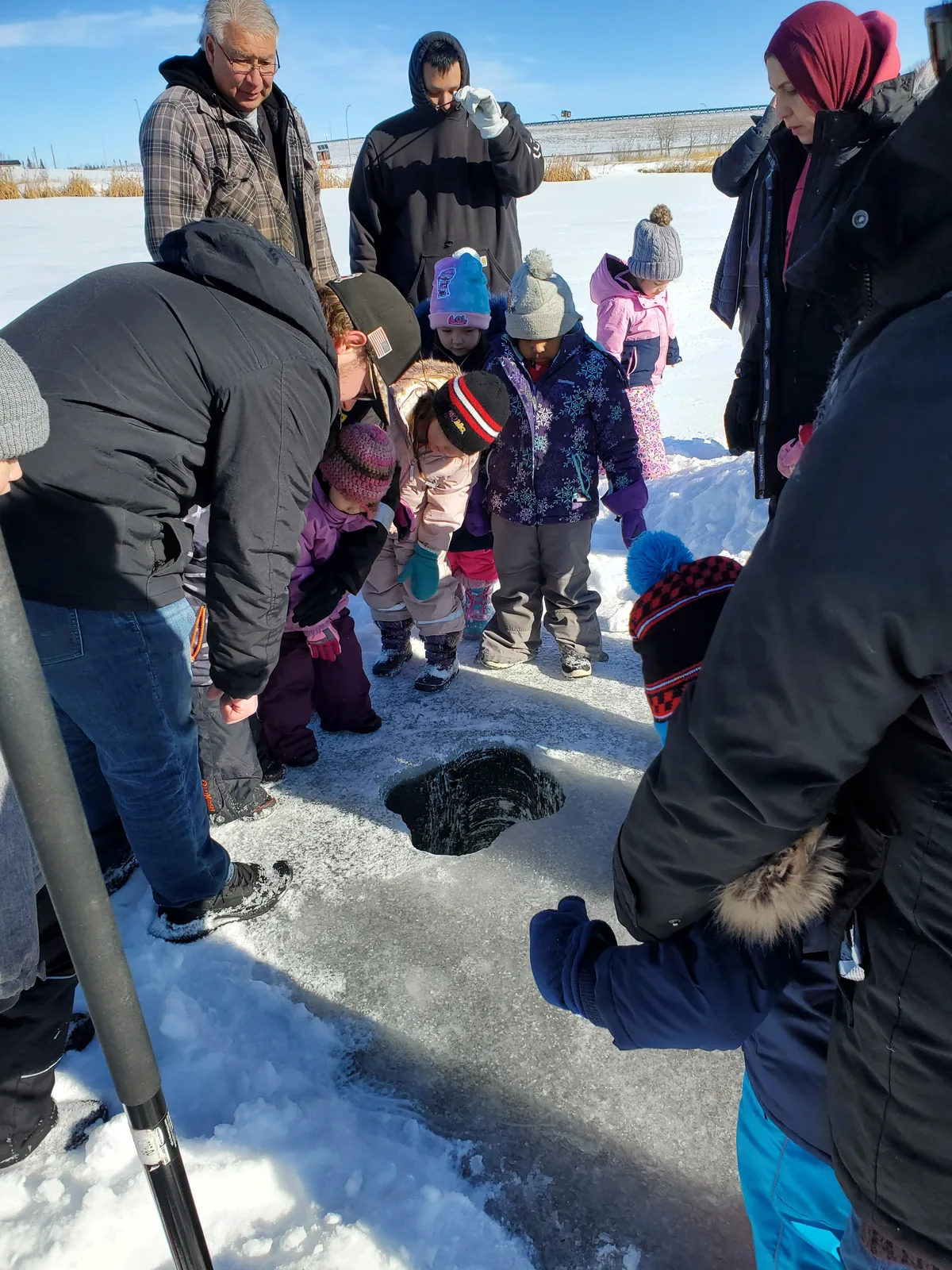 Children and adults gathered around a hole in the ice, a rod resting beside it.