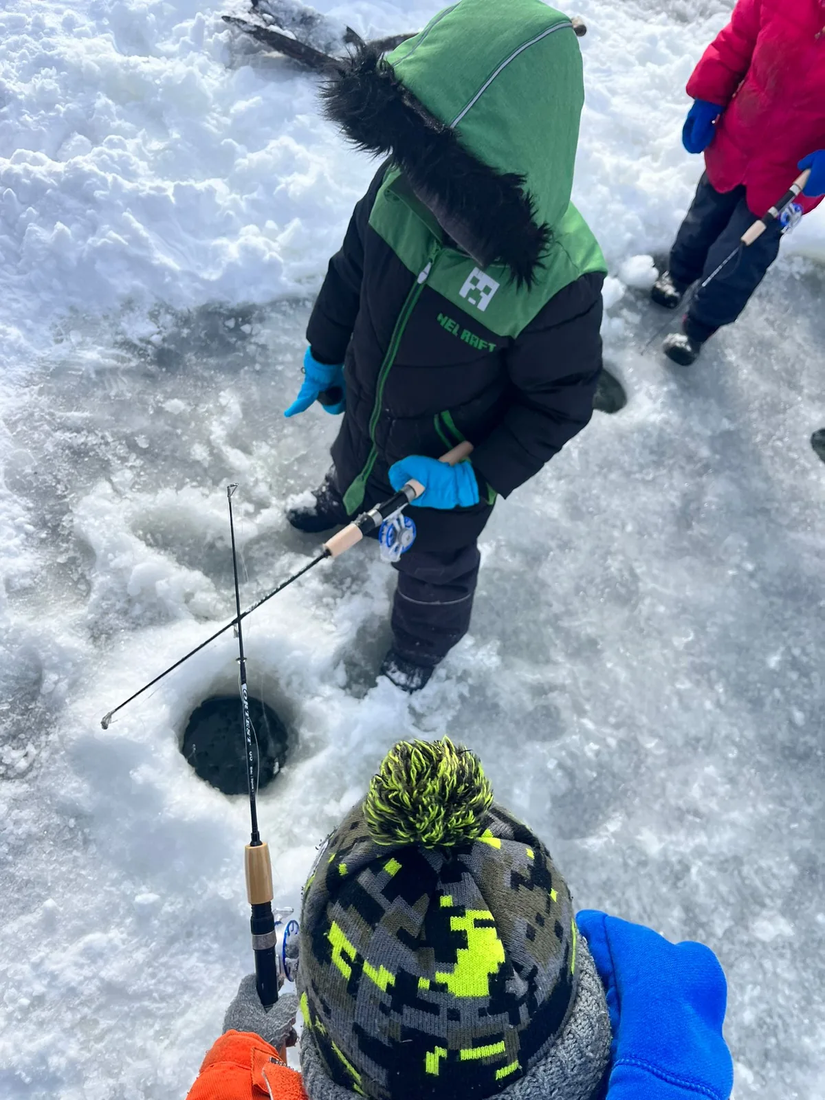 A child in a green hooded snowsuit holding a rod over a hole in the ice, seen from above.