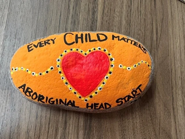 A painted stone in orange with a red heart at its centre, lettered EVERY CHILD MATTERS and ABORIGINAL HEAD START around the edge.