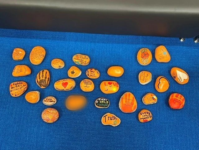 Around thirty painted orange stones laid out on a blue display board, carrying hearts, handprints, flowers, Cree words and messages.