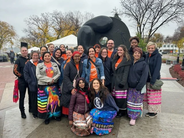 Staff gathered in front of a large black sculpture, many in orange shirts and ribbon skirts.