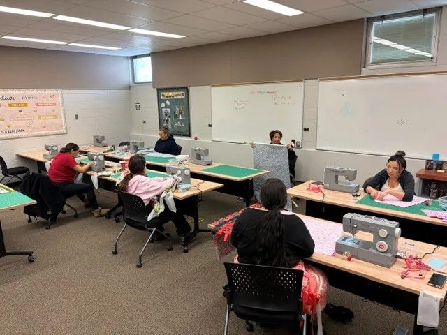 The class in progress, seven people spread across the room at their own machines, one holding up a length of dark fabric to check it.
