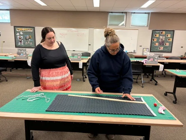 Two women measuring a length of navy patterned fabric on a cutting mat with a long wooden yardstick.