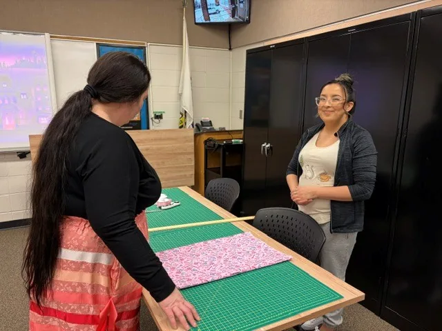 Two women at a cutting table, a piece of pink floral fabric laid out on the green mat between them.