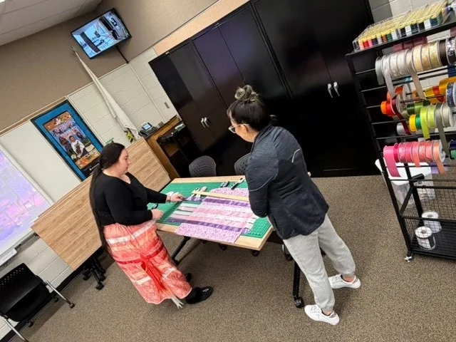 Two women at the cutting table laying out pale ribbon strips, a rack of ribbon spools in every colour beside them.