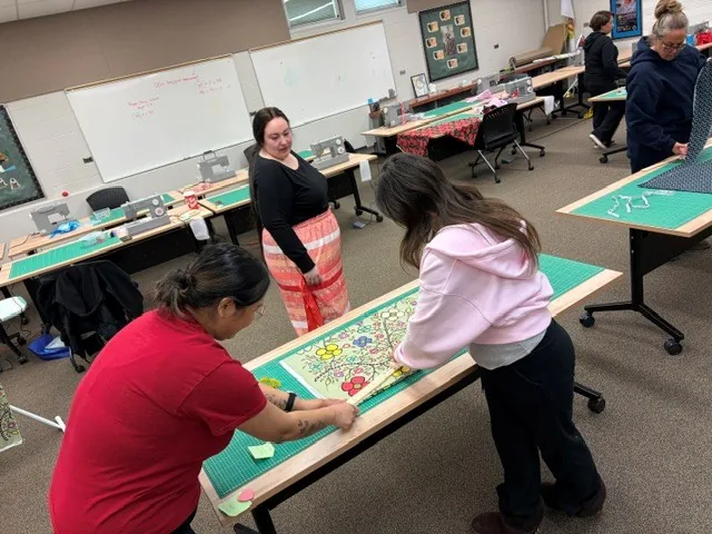 Several people gathered around a long table, pinning and marking the green floral panel before sewing.