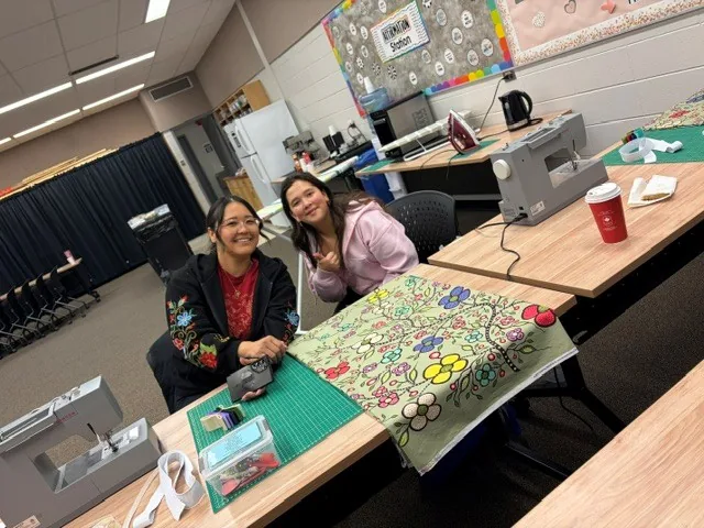 Two women seated at sewing machines, smiling at the camera, a green floral skirt panel spread out in front of them.