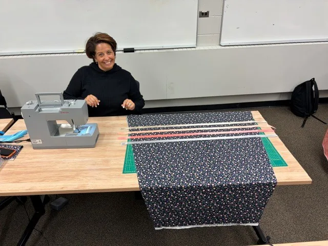 A woman at her machine with navy fabric laid out, rows of pink and cream ribbon already pinned across it.