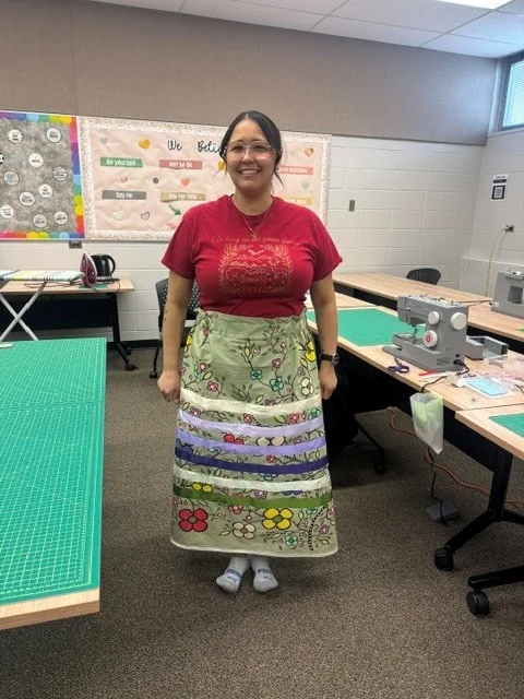 A woman standing in the classroom wearing her finished skirt, green with floral print and bands of ribbon around the hem.