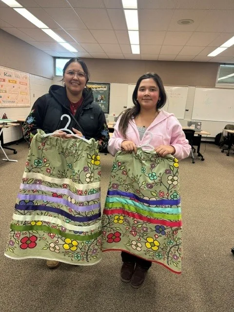 Two people holding up their finished green floral ribbon skirts on hangers, side by side.