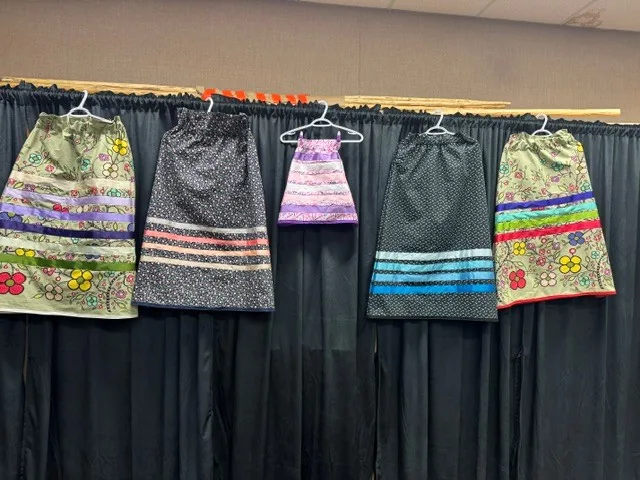 Five finished ribbon skirts hanging in a row on a rod, each with its own fabric and its own bands of ribbon.