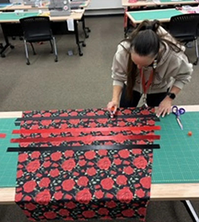 A woman measuring ribbon onto red floral fabric at a cutting table in the classroom.