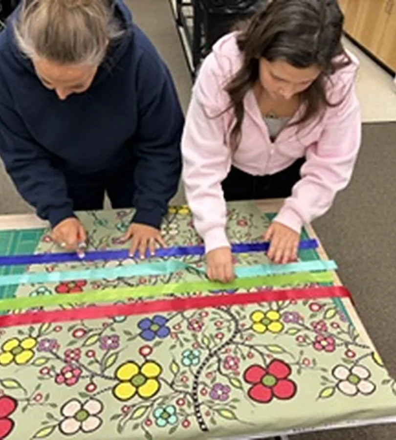Two women laying coloured ribbon across a length of green floral fabric on a cutting mat.