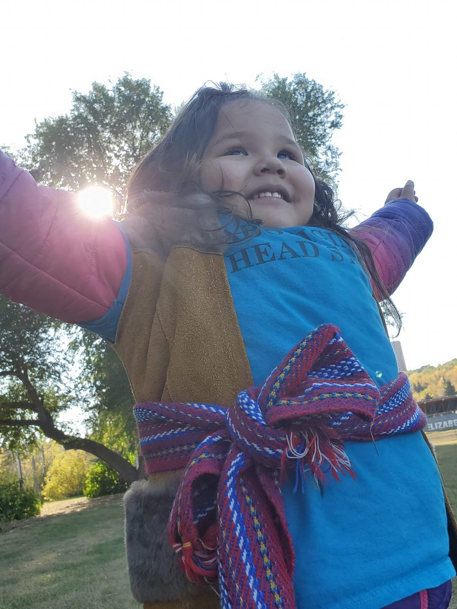 A child in a hide vest and woven sash standing outdoors with arms outstretched, smiling into the late afternoon sun.